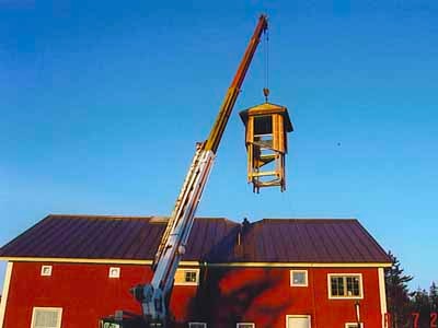 Crane setting cupola onto red Garner barn roof at dusk